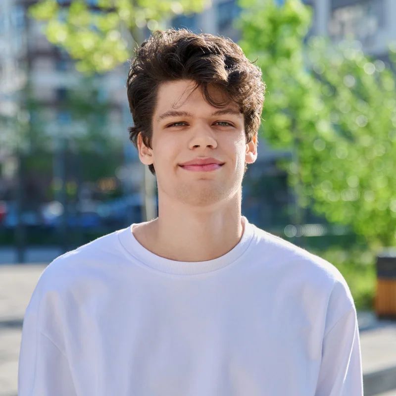 Young man with curly brown hair in a white sweatshirt smiling outdoors with green trees behind