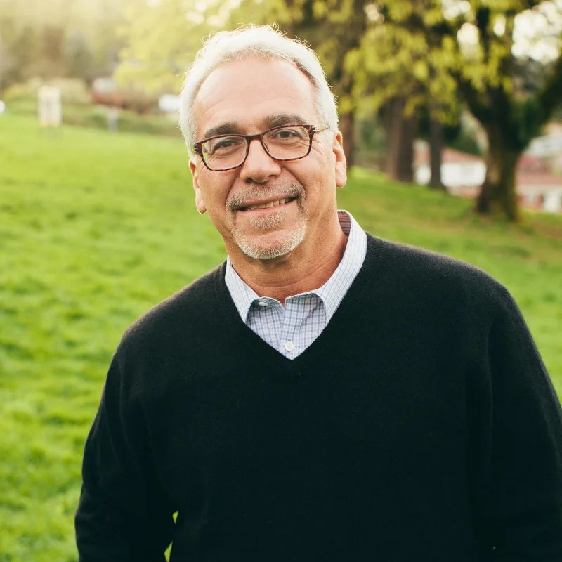 Smiling older man with gray hair and glasses in a dark sweater standing in a sunlit grassy park