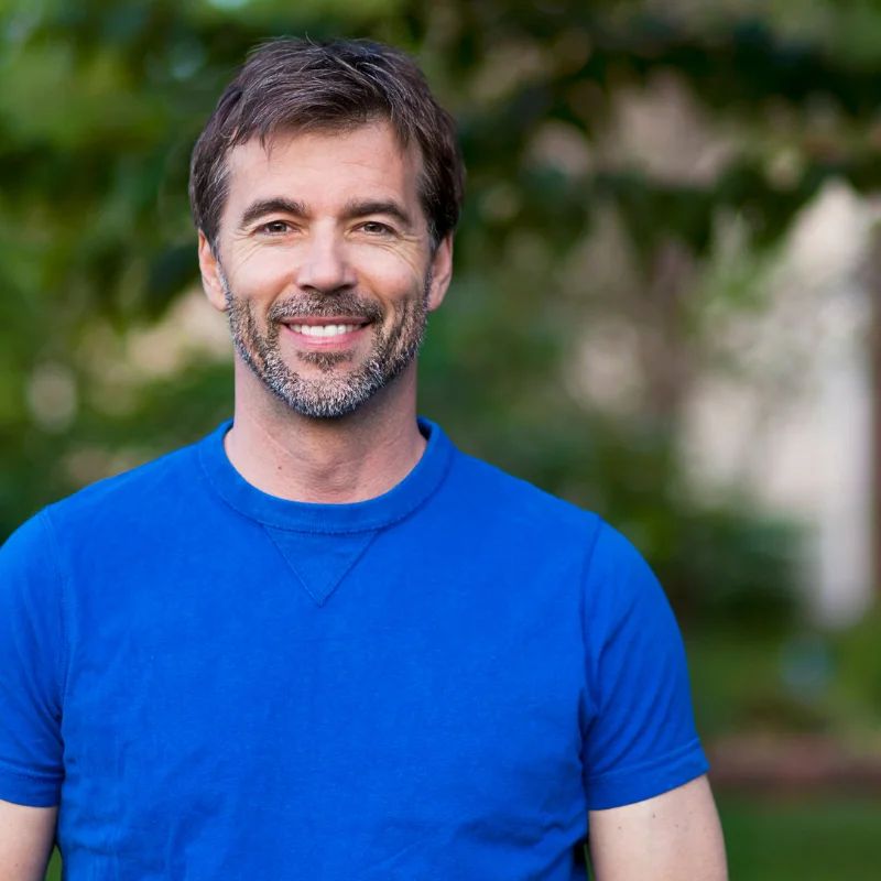Smiling bearded man in a blue t-shirt standing outdoors in front of greenery