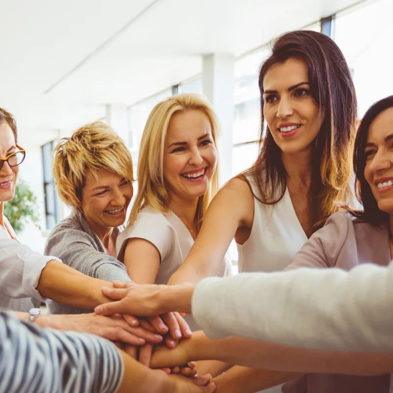 Group of smiling women stacking their hands together in the center of a sunlit room