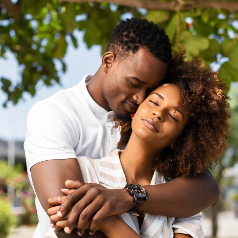 Couple embracing outdoors in sunlight, the woman resting against the man's chest