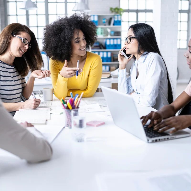 Four women collaborating around a table with a laptop and papers in a bright office