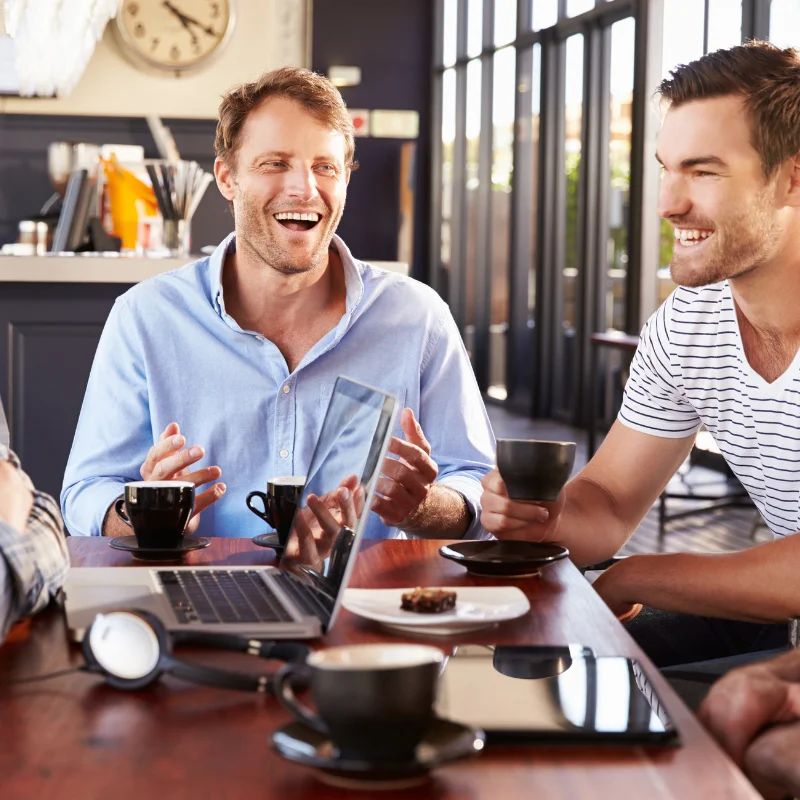 Two men laughing across a wood cafe table with espresso cups and an open laptop