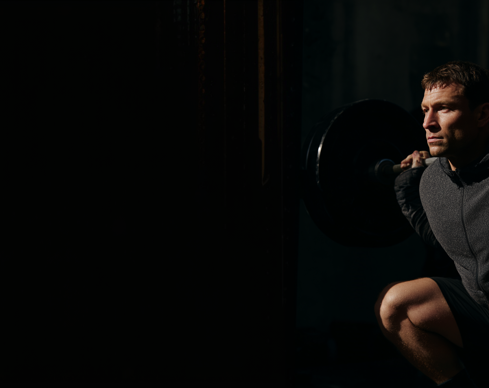 Athletic man crouched with a loaded barbell in a dark gym, lit from the side