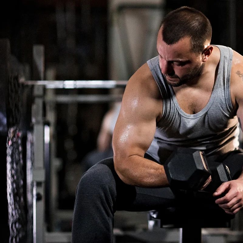 Muscular man in a gray tank top doing a seated dumbbell curl in a dim gym