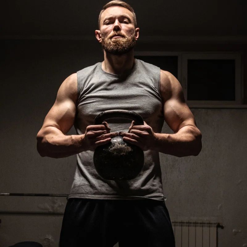 Muscular bearded man holding a kettlebell at his chest in a dim gym