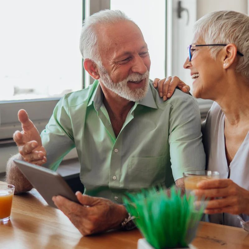 Older couple laughing together at a table while looking at a tablet